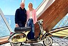 Ein Mann in dunklem Kapuzenpulli und eine Frau in rosafarbenem Jackett und Jeans hinter einer goldfarbenen Vespa. Durch das Fenster ist das blaue Meer zu sehen