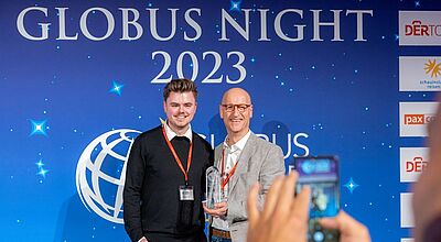 Zwei Männer stehen lächelnd vor einer blauen Wand mit der Aufschrift „GLOBUS NIGHT 2023“, einer davon hält einen Glaspokal in der Hand. Beide tragen rote Veranstaltungsausweise um den Hals. Im Vordergrund wird die Szene von einer Person mit Smartphone fotografiert.