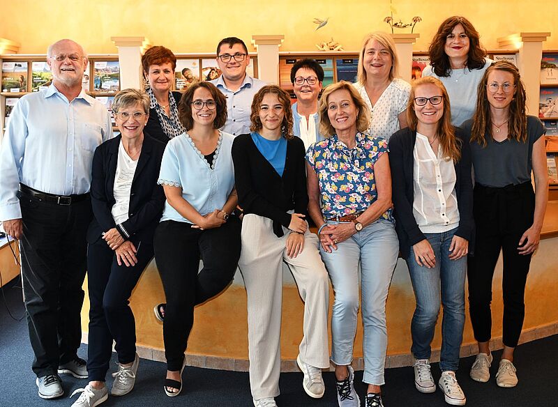 Gruppenfoto von zwölf lächelnden Personen, die in zwei Reihen in einem Büro oder Empfangsbereich stehen und sitzen. Im Hintergrund sind Regale mit Prospekten zu sehen.