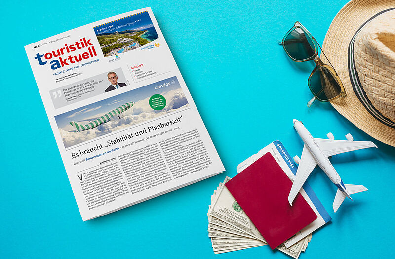 Ausgabe der Fachzeitschrift touristik aktuell liegt auf türkisfarbenem Untergrund, daneben Strohhut, Sonnenbrille, Reisepass und Dollarnoten