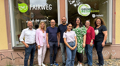 Eine Gruppe von acht Personen steht lächelnd vor einem Schaufenster mit den Logos Fairweg und Grünes Reisebüro. Die Menschen posieren gemeinsam auf einem gepflasterten Gehweg.