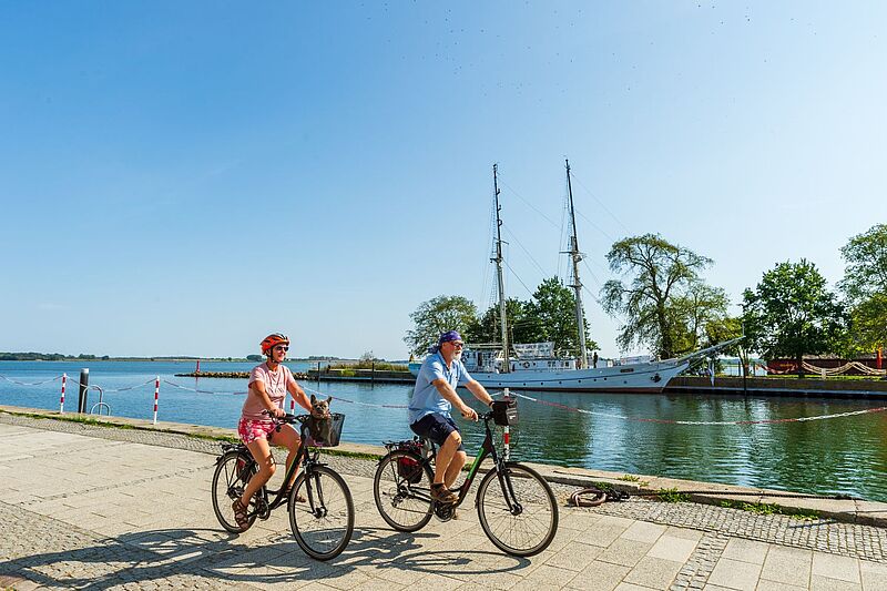 Eine neue Route hat der Veranstalter Mecklenburger Radtour auch für Usedom ausgearbeitet. Zwei Personen in Sommerkleidung fahren bei sonnigem Wetter mit Fahrrädern an einem Hafen entlang, im Wasser liegt ein Segelschiff.