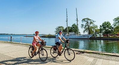 Eine neue Route hat der Veranstalter Mecklenburger Radtour auch für Usedom ausgearbeitet. Zwei Personen in Sommerkleidung fahren bei sonnigem Wetter mit Fahrrädern an einem Hafen entlang, im Wasser liegt ein Segelschiff.