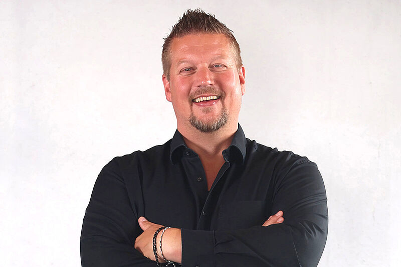 Auf dem Bild ist ein Mann in einem eleganten, schwarzen Hemd mit verschränkten Armen vor einem hellen, minimalistischen Hintergrund zu sehen. Das Portrait vermittelt Selbstbewusstsein und Seriosität.