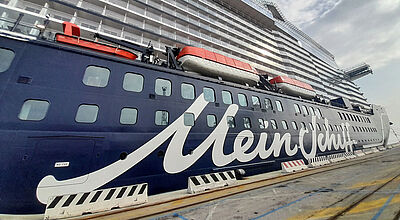 Seitenansicht eines großen Kreuzfahrtschiffes an der Pier. Auf dem dunkelblauen Rumpf prangt groß in Schreibschrift der Name Mein Schiff.