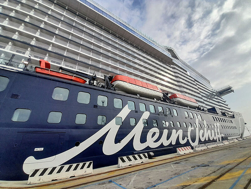 Seitenansicht eines großen Kreuzfahrtschiffes an der Pier. Auf dem dunkelblauen Rumpf prangt groß in Schreibschrift der Name Mein Schiff.