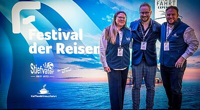 Glücklich über ein erfolgreiches Event (von links): Die Reisebüro-Inhaber Anna Lena (links) und Aron Stiefvater mit Philipp Holzinger als Vertreter des Top-Sponsors MSC Cruises. Drei Personen stehen lächelnd vor einem blau beleuchteten Messe- oder Event-Hintergrund mit der Aufschrift Festival der Reisen. Sie tragen Namensschilder und dunkelblaue Westen. Hinter ihnen ist ein Meer- und Kreuzfahrtmotiv zu sehen