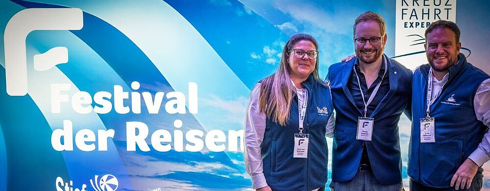 Drei Personen stehen lächelnd vor einem blau beleuchteten Messe- oder Event-Hintergrund mit der Aufschrift Festival der Reisen. Sie tragen Namensschilder und dunkelblaue Westen. Hinter ihnen ist ein Meer- und Kreuzfahrtmotiv zu sehen