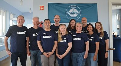 Fünf Frauen und fünf Männer lächeln in die Kamera. Alle tragen ein blaues T-Shirt auf dem Moin steht
