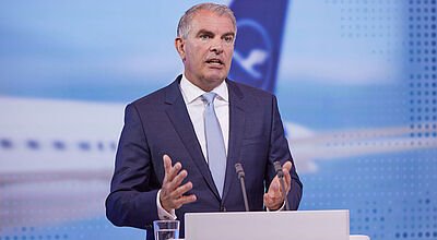 Ein Konzern-Manager mittleren Alters in dunklem Anzug, weißem Hemd und blauer Krawatte steht redend und gestikulierend hinter einem Rednerpult. Im Hintergrund ist unscharf auf einer Leinwand eine Heckflosse eines Lufthansa-Flugzeugs zu sehen.