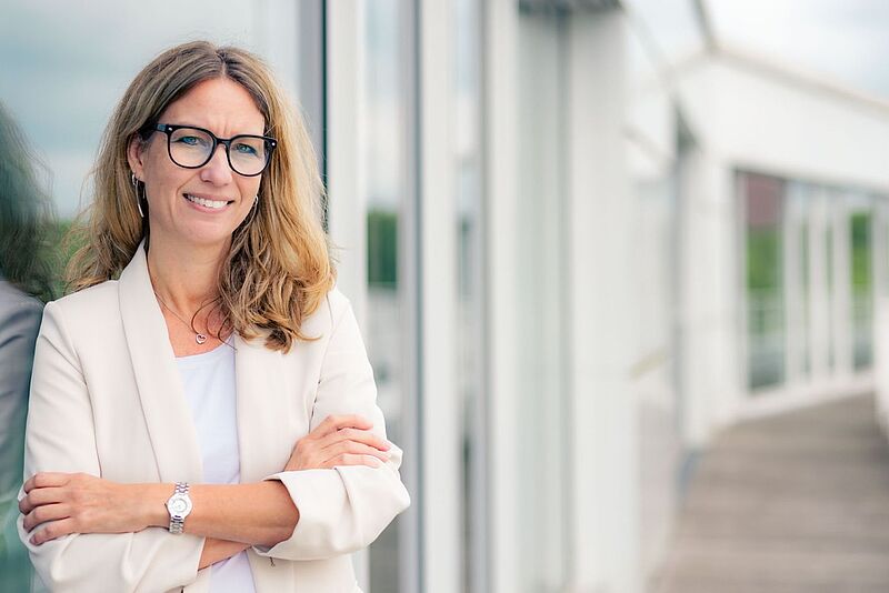 Eine Frau mit langen, welligen, blonden Haaren und schwarzer Brille steht mit verschränkten Armen vor einer Glasfassade. Sie trägt einen cremefarbenen Blazer über einem weißen Oberteil und eine silberne Armbanduhr. Sie lächelt freundlich und blickt direkt in die Kamera. Der Hintergrund ist modern mit großen Glasfenstern und einem Holzsteg.