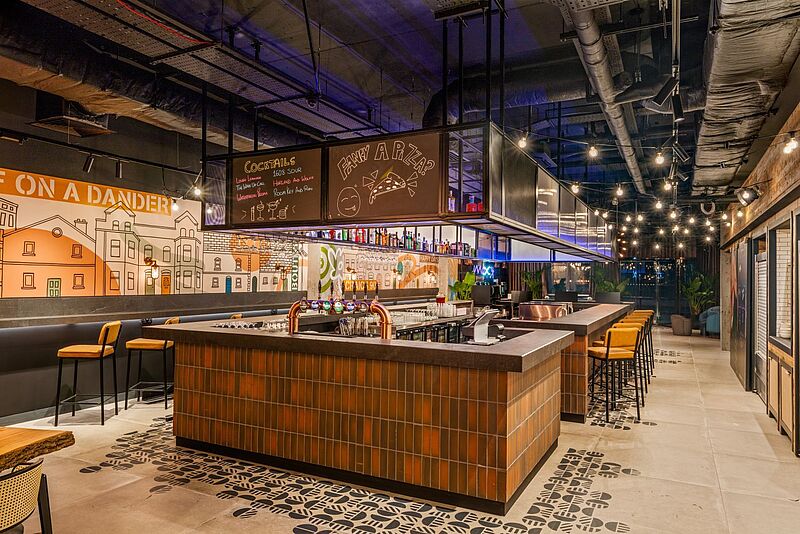 An dieser Bar im Moxy Belfast City checken die Gäste ein und bekommen ihren Welcome Drink Stylische Hotelbar im Moxy Belfast City, Nordirland – modernes Interior mit beleuchtetem Tresen, farbenfroher Wandgestaltung, gemütlichen Sitzplätzen und trendiger Atmosphäre für Gäste und Städtereisende.