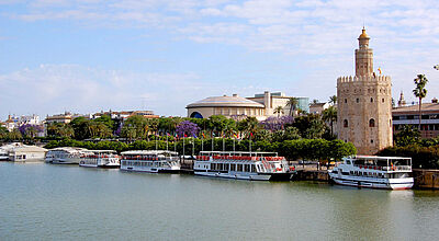 Der Besuch von Sevilla darf bei keiner der neuen Rundreisen fehlen. Foto: ras Blick auf den Guadalquivir-Fluss in Sevilla mit mehreren Ausflugsbooten am Ufer, im Hintergrund die von Palmen und blühenden Bäumen umgebene Goldene Turm (Torre del Oro), ein historisches Wahrzeichen aus maurischer Zeit, daneben moderne Gebäude unter leicht bewölktem Himmel.