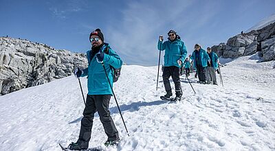 Schneeschuh-Wanderungen sind künftig fester Bestandteil des Ausflugsprogramms bei Antarktis-Reisen Eine kleine Gruppe von Antarktis-Touristen in dunklen Schneehosen und einheitlichen dunkelblauen Anoraks unternimmt eine Schneeschuhwanderung. Die Teilnehmer schützen sich mit Sonnenbrillen gegen die Sonne und sichern sich mit Walking-Stöcken im Schnee.