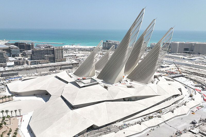 Futuristisches Gebäude mit geschwungenen Stahlbögen vor der Skyline von Abu Dhabi und Meerblick. Weiße geometrische Struktur mit kalligrafischen Fenstern.