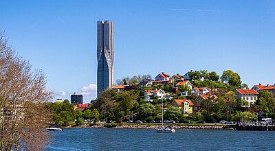 Foto von einem Wolkenkratzer unter ein paar Häusern vor blauem Himmel. Im Vordergrund ist Wasser zu sehen.