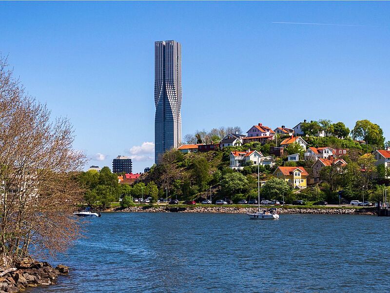 Foto von einem Wolkenkratzer unter ein paar Häusern vor blauem Himmel. Im Vordergrund ist Wasser zu sehen.