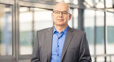 Ein glatzköpfiger Mann mittleren Alters mit Brille steht in einem modernen Bürogebäude. Er trägt ein graues Sakko über einem blauen Hemd und blickt selbstbewusst in die Kamera. Im Hintergrund sind große Fensterfronten mit Glasrahmen und unscharfe Außenansichten zu sehen.