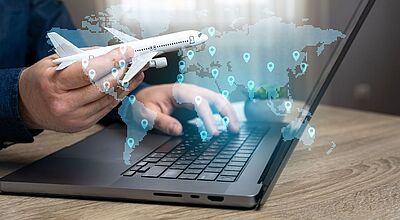 Ein Mann sitzt an einem schwarzen Laptop und hält in der Hand ein weißes Modellflugzeug. Abgesoftet zu sehen sind die Umrisse verschiedener Kontinente sowie Zielgebiete.