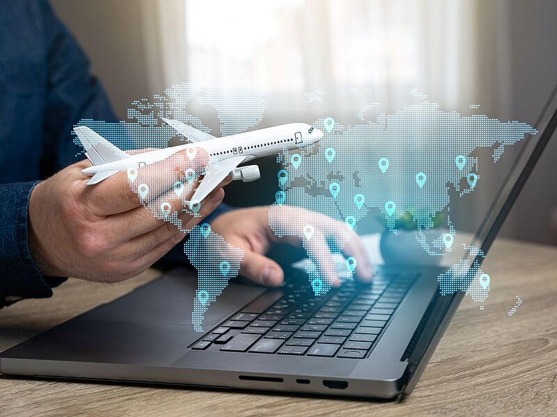Ein Mann sitzt an einem schwarzen Laptop und hält in der Hand ein weißes Modellflugzeug. Abgesoftet zu sehen sind die Umrisse verschiedener Kontinente sowie Zielgebiete.
