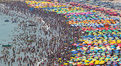Luftaufnahme eines überfüllten Strandes: Tausende Menschen im Wasser und am Ufer, dicht an dicht gereihte bunte Sonnenschirme am Sandstrand.