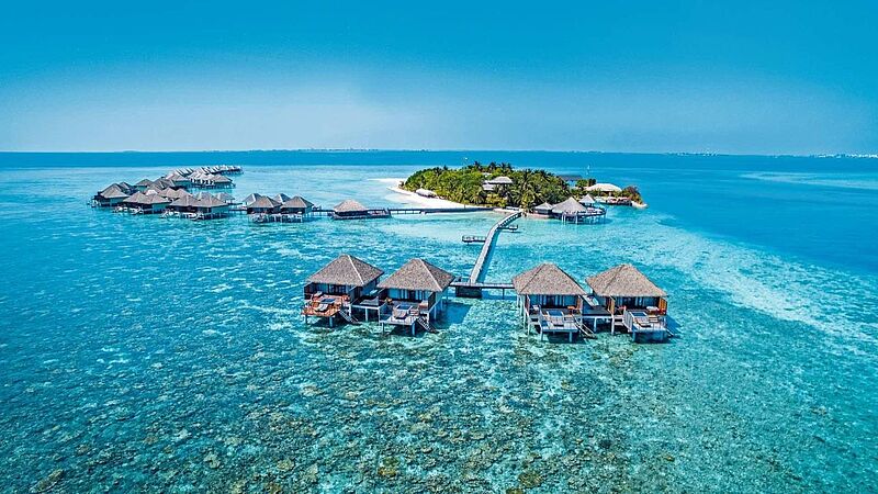 Luftaufnahme eines tropischen Inselresorts mit Wasserbungalows auf türkisblauem Meer, die durch Holzstege mit einer kleinen, üppig bewachsenen Insel mit weißen Sandstränden verbunden sind. Der Himmel ist klar und blau, das Wasser wirkt flach und kristallklar.