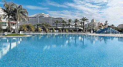 Großer, einladender Hotelpool mit klarem, blauem Wasser vor einer modernen Hotelanlage und Palmen unter blauem Himmel; im Hintergrund ein verspielter Wasserpark und ein prachtvolles Gebäude mit Türmen im orientalischen Stil.