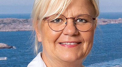Porträtfoto einer blonden Dame mit Brille und Business-Outfit. Sie trägt ein kariertes Sakko auf einer weißen Bluse und lächelt in die Kamera. Im Hintergrund ist eine Luftaufnahme eines Seehafens zu erkennen.