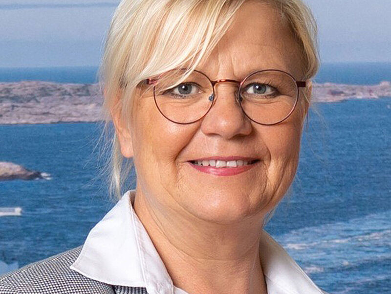 Porträtfoto einer blonden Dame mit Brille und Business-Outfit. Sie trägt ein kariertes Sakko auf einer weißen Bluse und lächelt in die Kamera. Im Hintergrund ist eine Luftaufnahme eines Seehafens zu erkennen.