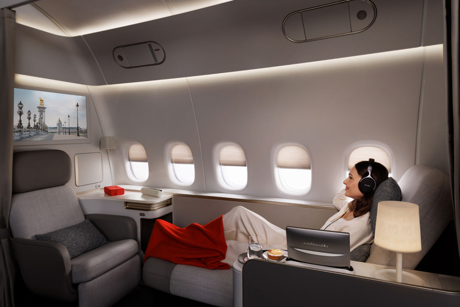 First-Class-Flugzeugkabine von Air France mit grauer Polsterung, roter Decke, Laptop und Kopfhörer. Monitor zeigt Pariser Brückenszene. Ambiente-Beleuchtung und fünf Fenster.