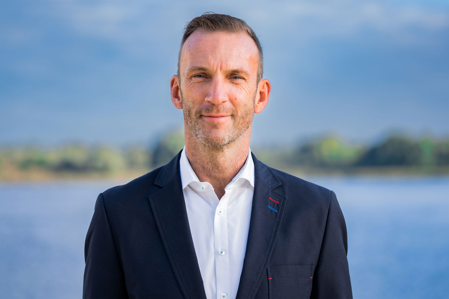 Der Vertriebschef einer Reederei Mitte vierzig steht in dunkelblauem Sakko freundlich lächelnd vor der Kamera. Er hat kurze braune Haare und einen Bartansatz.