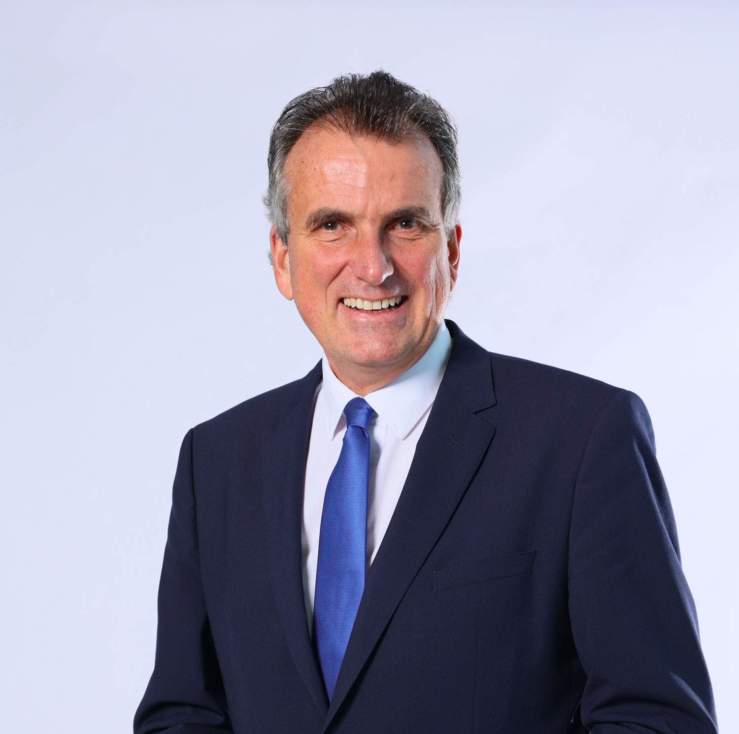 Porträtfoto eines älteren Reederei-Managers mit braun-grauem Haar, dunklem Anzug und blauer Krawatte. Er blickt lächelnd in die Kamera. 