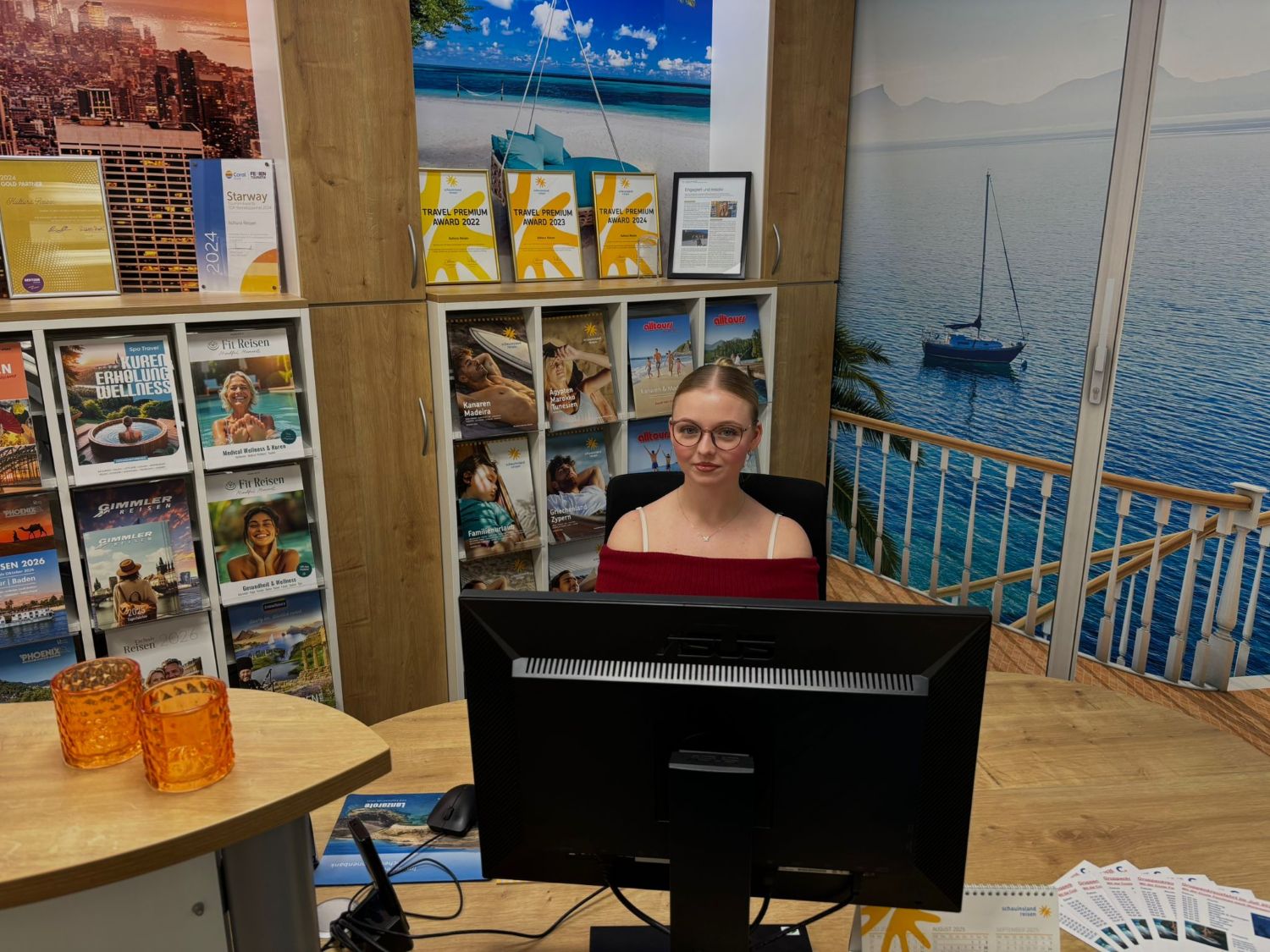 Eine junge Frau sitzt hinter einem schwarzen Monitor am Schreibtisch. Hinter dem Schreibtisch sind Kataloge zu sehen. Sie lächelt in die Kamera