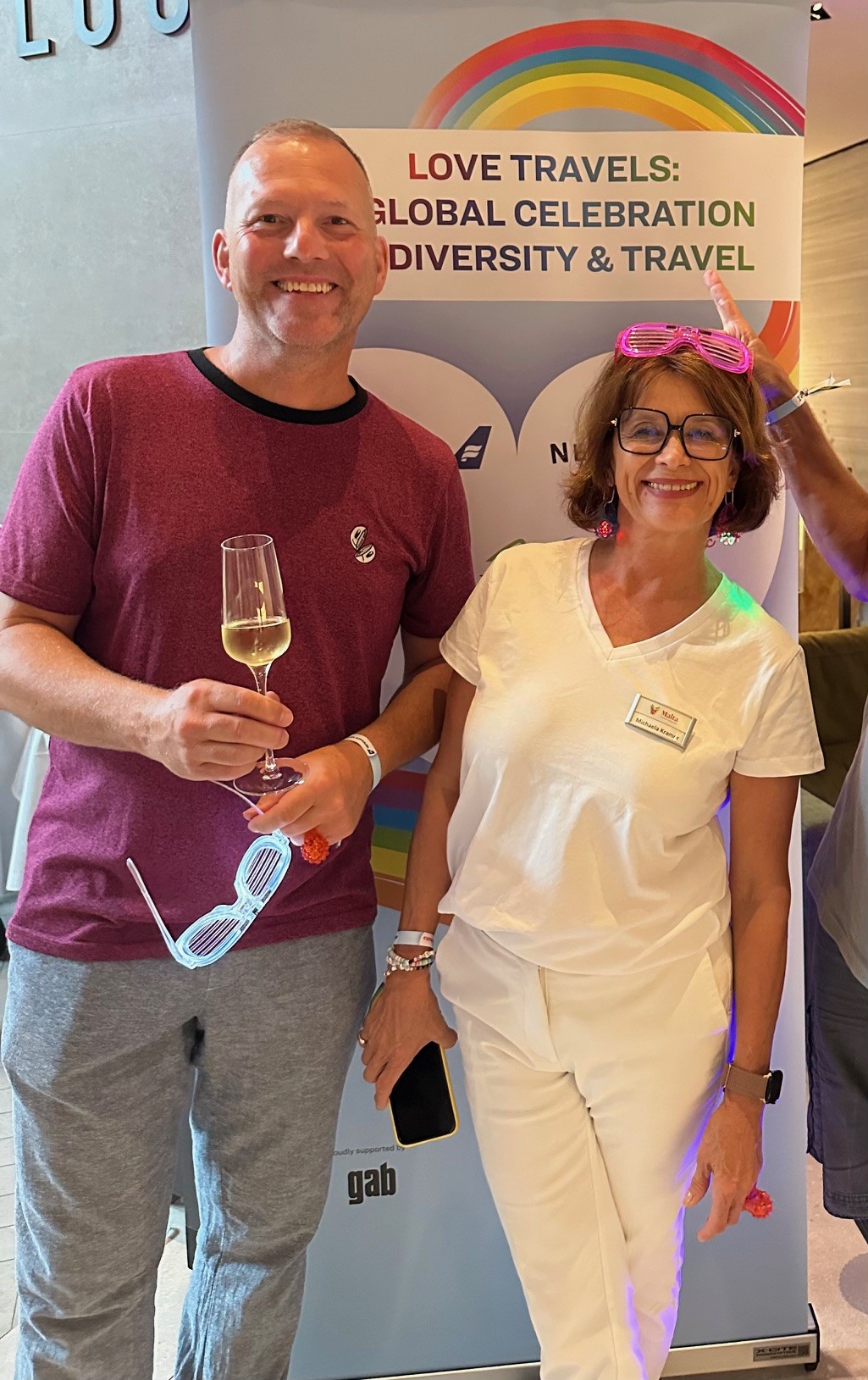 Zwei Personen posieren lächelnd vor einem Banner mit der Aufschrift „LOVE TRAVELS: GLOBAL CELEBRATION OF DIVERSITY & TRAVEL“ und einem Regenbogenmotiv. Die Frau trägt weiße Kleidung, auffällige pinke Accessoires und hält ein Smartphone, während der Mann ein Glas Sekt in der Hand hält und eine Sonnenbrille trägt. Die Atmosphäre wirkt entspannt und freundlich, passend zu einer Pride- oder Diversity-Veranstaltung.