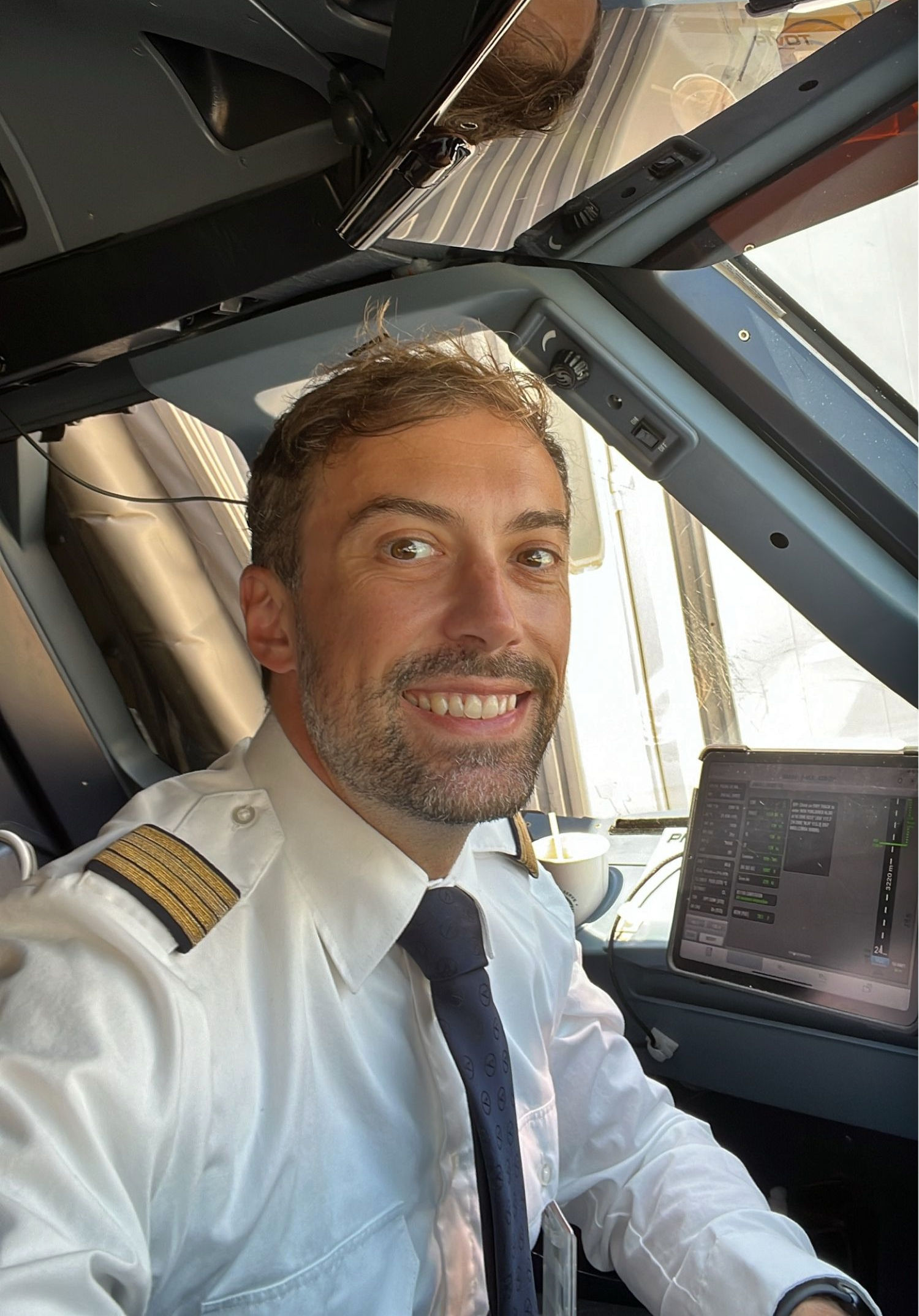 Ein Pilot in weißer Uniform mit Schulterabzeichen und dunkler Krawatte sitzt im Cockpit eines Flugzeugs und lächelt in die Kamera. Neben ihm ist ein Tablet mit Fluginformationen zu sehen.
