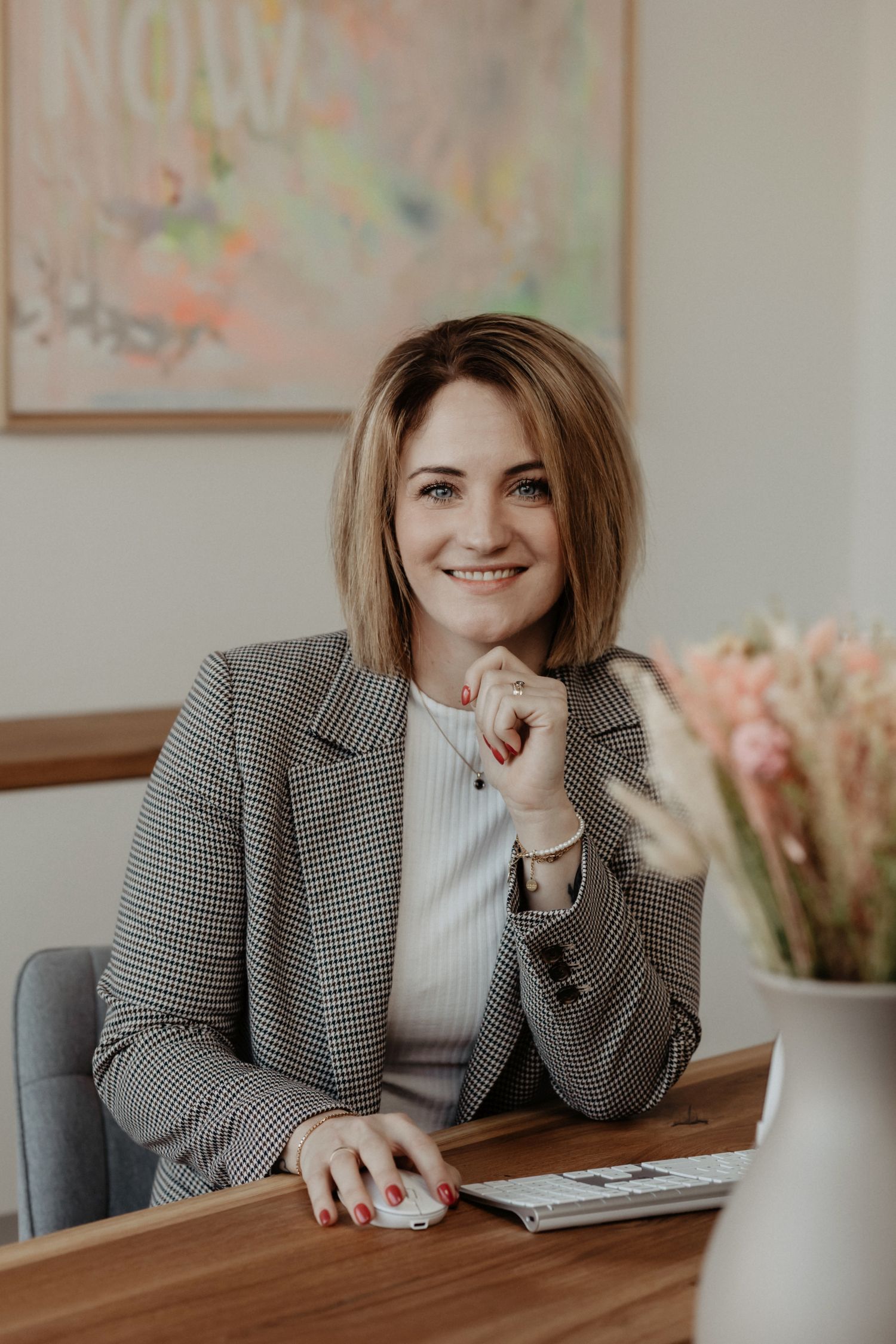 Frau mit schulterlangem blond-braunem Haar sitzt lächelnd an einem Schreibtisch. Sie trägt einen karierten Blazer und ein weißes Oberteil, hat eine Hand am Kinn und die andere auf einer Computermaus. Auf dem Tisch stehen eine Tastatur und eine Vase mit getrockneten Blumen.