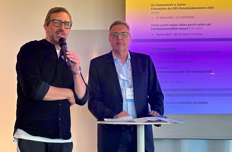 Zwei Männer stehen nebeneinander auf einer Bühne vor einer Präsentationsfolie. Der Mann links spricht in ein Mikrofon, trägt eine dunkle Hemdjacke und eine Brille. Der Mann rechts trägt einen dunklen Anzug mit hellblauem Hemd und steht an einem kleinen Stehtisch mit Papieren darauf. Hinter ihnen ist der Text einer Präsentation zum Thema Reisewirtschaft zu sehen.