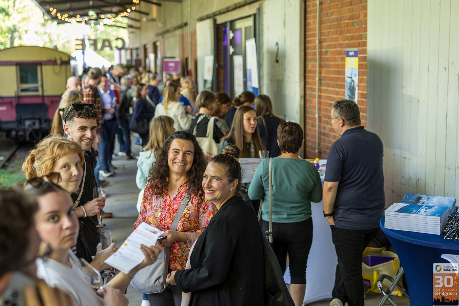Eine große Gruppe von Menschen besucht eine Outdoor-Messe beim Sommerstammtisch des Deutschen Reiserings. Die Veranstaltung findet an einem Bahnsteig mit historischem Waggon im Hintergrund statt. An mehreren Infoständen entlang einer Backsteinwand informieren sich Besucher über touristische Angebote. Einige Personen lächeln in die Kamera, andere sind in Gespräche vertieft. Im Vordergrund liegen Broschüren und Werbematerialien aus. In der rechten unteren Ecke ist das Logo des Deutschen Reiserings mit dem Hinweis auf das 30-jährige Jubiläum zu sehen. Die Stimmung ist lebendig und interessiert.