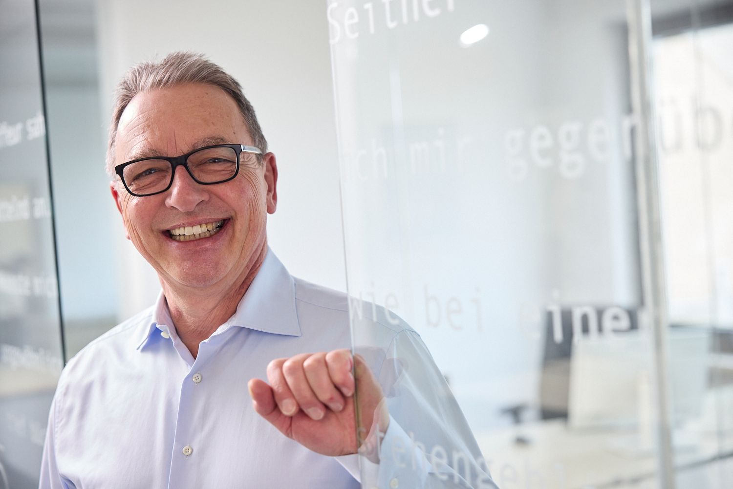 Ein älterer Mann mit grauen Haaren und Brille, es ist der Chef der Ziel GmbH Gerd Laatz, steht lächelnd in einem modernen Büro hinter einer Glaswand mit deutscher Schrift. Er trägt ein hellblaues Hemd und wirkt fröhlich und entspannt. Die Umgebung ist hell und professionell.