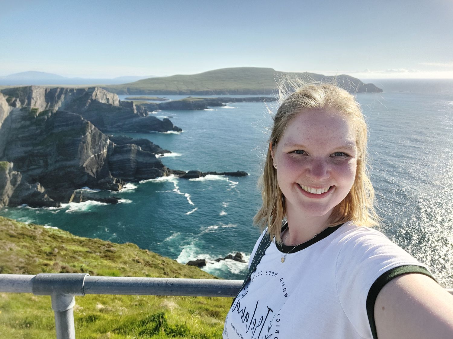 Eine blonde junge Frau lächelt in die Kamera macht ein Selfie vor einer spektakulären Kulisse aus schroffen grünen Felsen und blauem Meer.