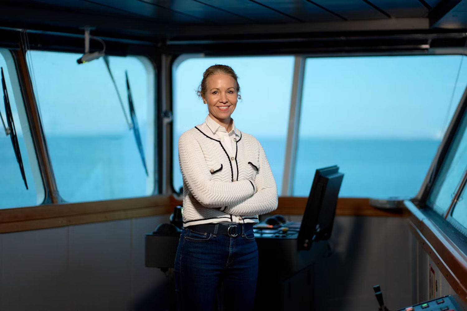 Die Chefin einer Reederei steht auf der Brücke eines Küstenkreuzfahrtschiffes und blickt lächelnd in die Kamera. Die Frau hat blondes Haar, trägt einen blau-weißen Wollpullover auf weißer Bluse sowie blaue Jeans.  