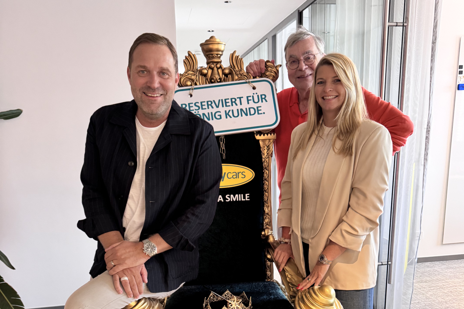 Drei Personen stehen und sitzen lächelnd vor einem goldverzierten Stuhl mit einem Schild, auf dem „Reserviert für König Kunde“ steht. Im Hintergrund ist ein Flur mit Glaswänden zu sehen.