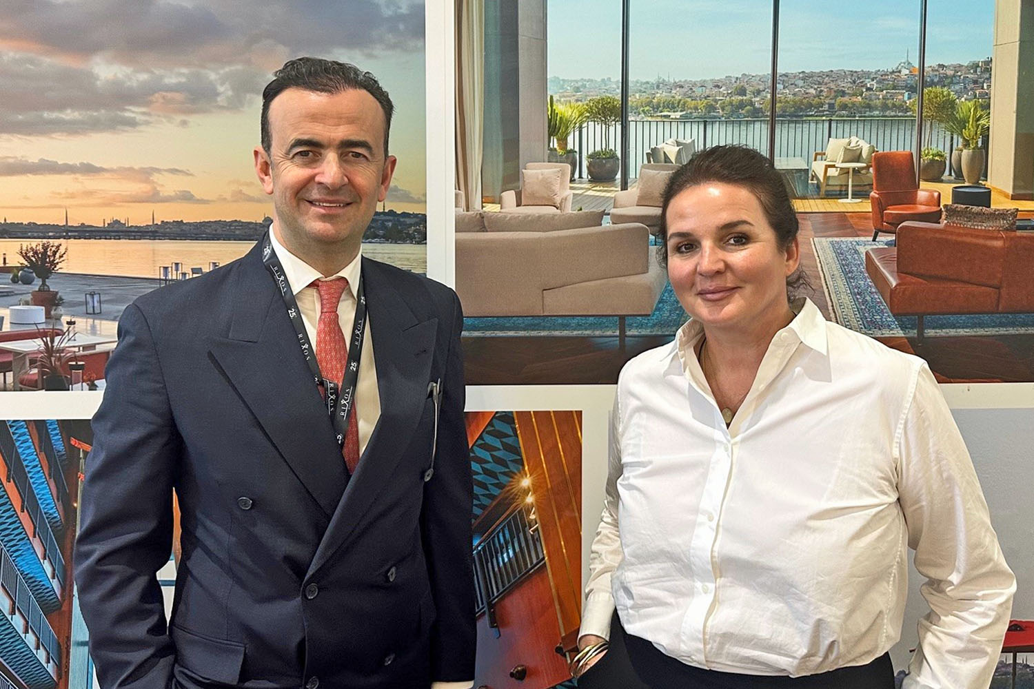 Ein Mann und eine Frau stehen vor einer großen Werbewand mit Bildern eines luxuriösen Resorts in Istanbul. Der Mann trägt einen dunklen Anzug mit roter Krawatte und hat ein Namensschild um den Hals. Er hat kurze, dunkle Haare und lächelt leicht. Die Frau trägt eine weiße Bluse und schwarze Hose, hat braune Haare locker zurückgebunden und lächelt freundlich. Die Werbewand zeigt stilvolle Innenräume mit großen Fenstern und Blick auf das Wasser sowie eine elegante Außenterrasse bei Sonnenuntergang.