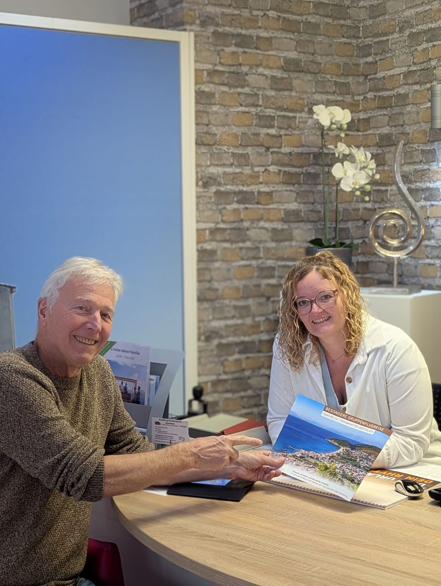 Eine Reiseberaterin und ein älterer Mann sitzen an einem Schreibtisch und betrachten gemeinsam einen Reisekatalog. Beide lächeln, während sie über ein Angebot sprechen. Im Hintergrund sind eine Backsteinwand, Deko-Elemente und Informationsmaterialien zu sehen.