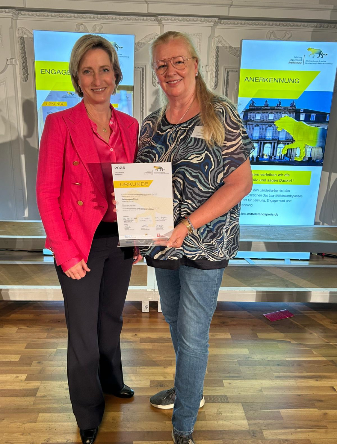 Die Auszeichnung übergab Baden-Württembergs Wirtschaftsministerin Nicole Hoffmeister-Krau Zwei Frauen stehen nebeneinander auf einer Bühne in einem festlichen Saal. Die Frau links trägt einen pinken Blazer und die Frau rechts, mit langen blonden Haaren und Brille, hält eine Urkunde in den Händen. Im Hintergrund sind Bildschirme mit dem Logo des LEA-Mittelstandspreises für soziale Verantwortung in Baden-Württemberg sowie die Worte „Engagement“ und „Anerkennung“ zu sehen. Die Szene zeigt eine offizielle Preisverleihung.