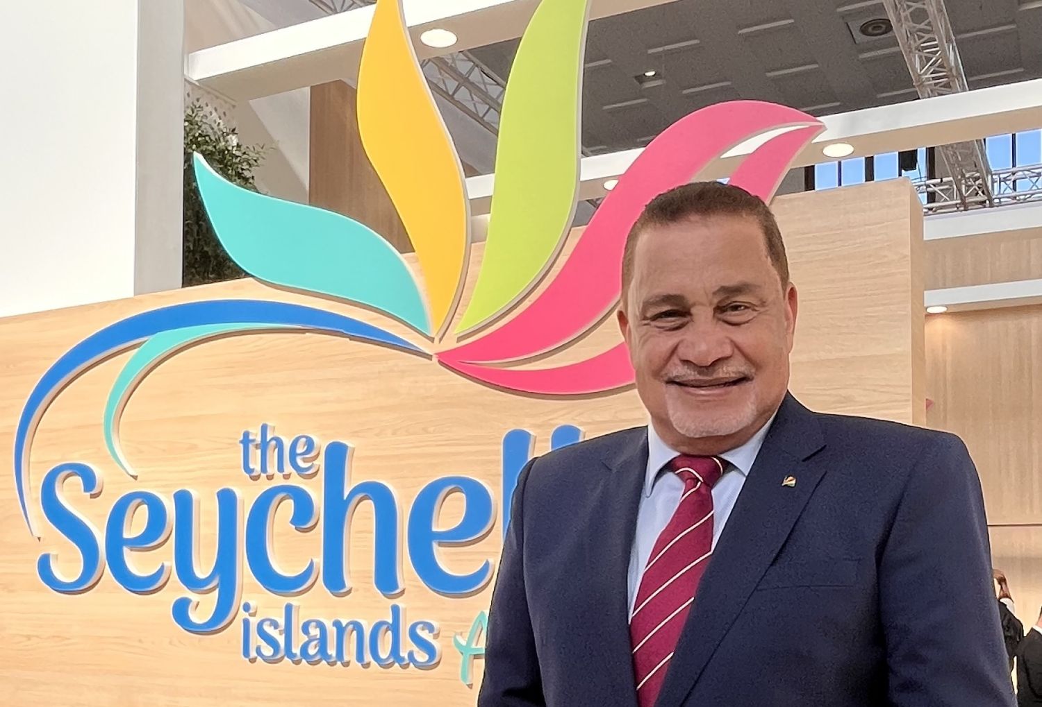 Geschäftsmann vor Seychellen-Logo mit bunten, geschwungenen Blättern in Türkis, Gelb, Hellgrün und Pink auf hellem Holzhintergrund.