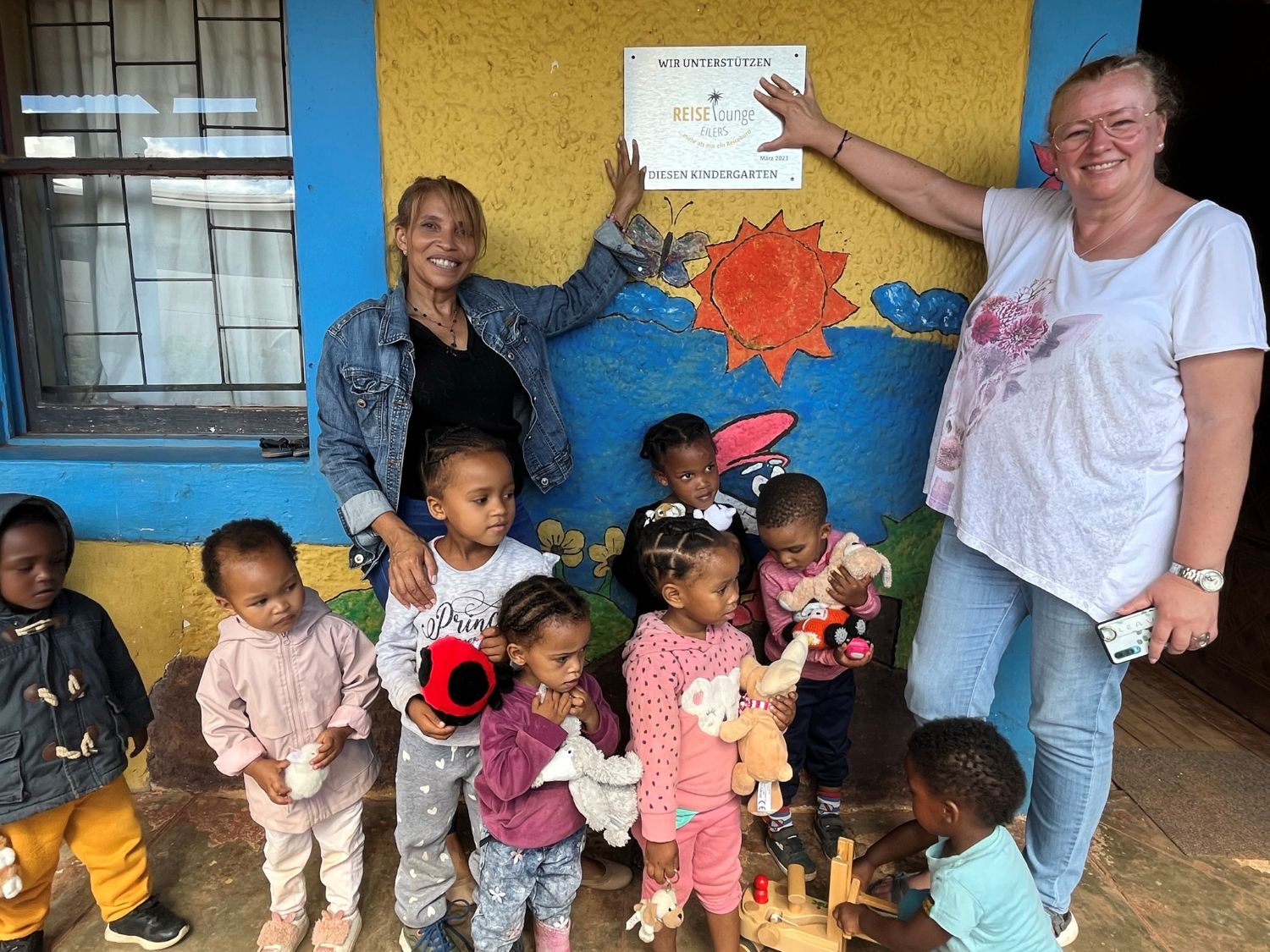 Eine Plakette am Schulgebäude zeigt, dass das Projekt von der Reiselounge Eilers unterstützt wird. Im Bild Annette Eilers und Anneline von Rooyen Besucher und Kinder in einem Kindergarten mit gelben Wänden, bunten Wandmalereien und Spielsachen. Ein Schild zeigt die Unterstützung durch die Reiselounge.