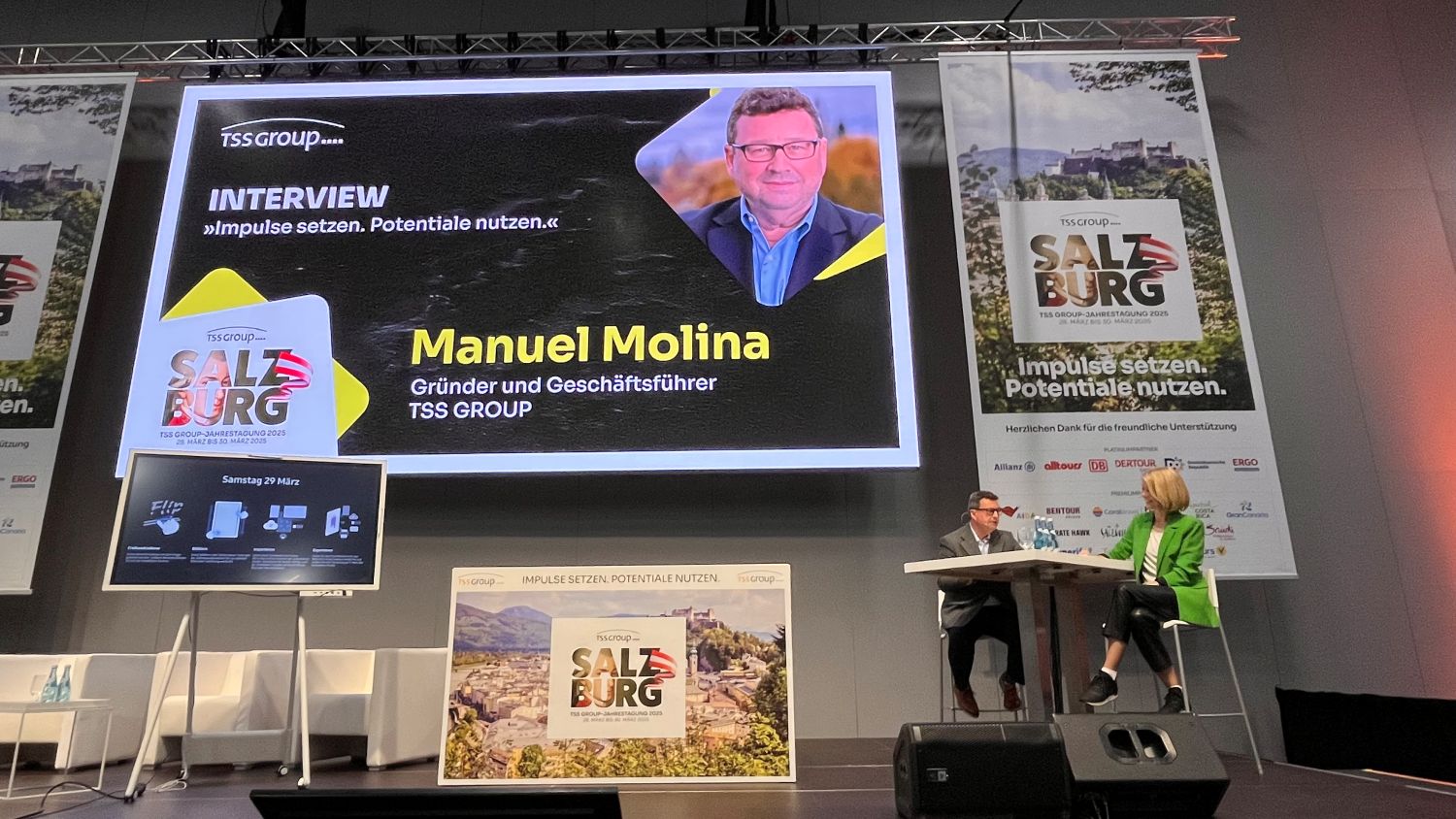Eine Bühne bei der TSS-Jahrestagung 2025 in Salzburg. Auf einer großen Leinwand ist ein Interview mit Manuel Molina, Gründer und Geschäftsführer der TSS Group, angekündigt. Der Slogan „Impulse setzen. Potentiale nutzen.“ ist mehrfach zu sehen. Zwei Personen sitzen auf der Bühne an einem Tisch und sprechen miteinander. Im Hintergrund sind Banner mit dem Veranstaltungslogo und den Namen verschiedener Sponsoren zu erkennen.