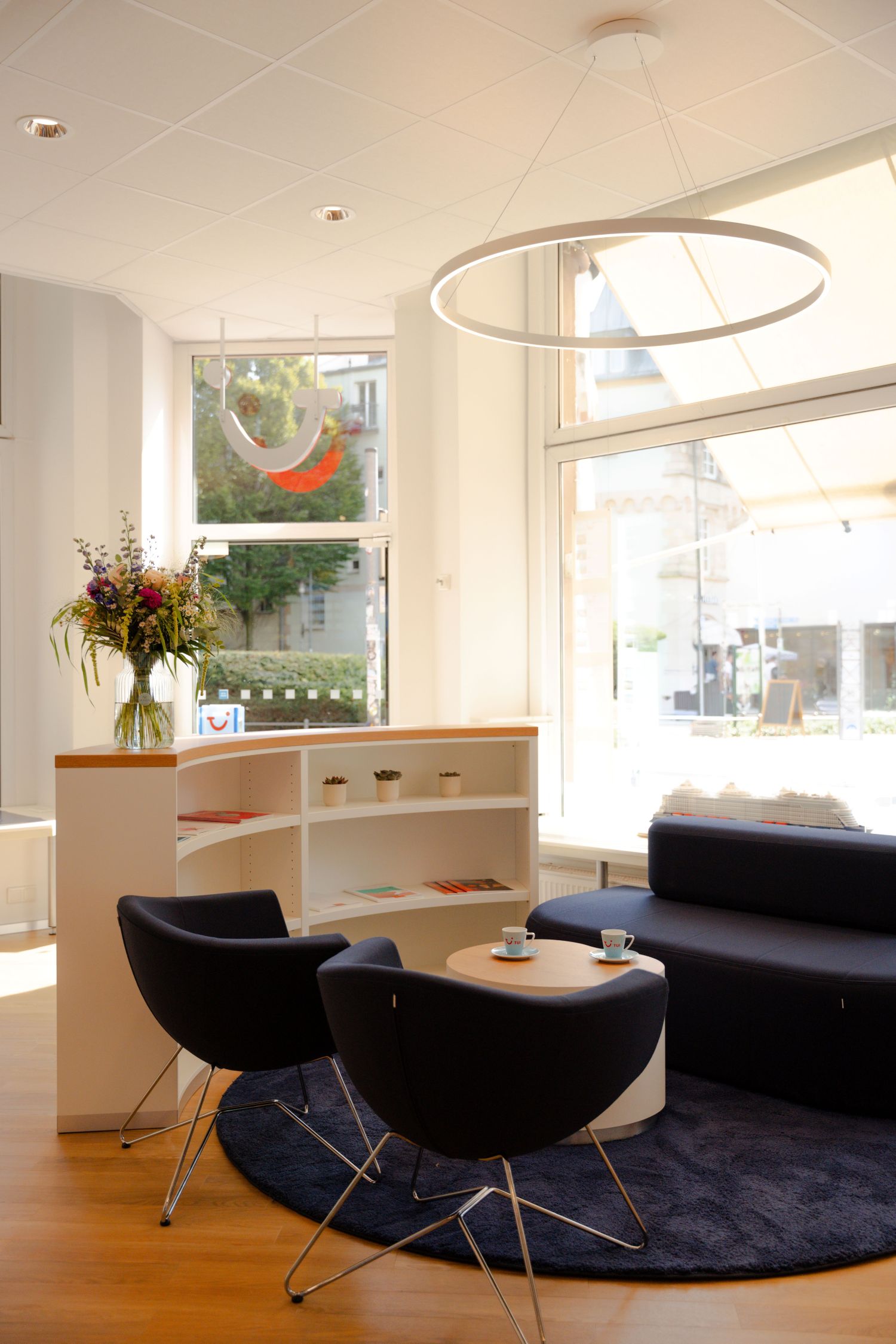 Der Eingangsbereich verfügt nun über einen großzügigen Empfangstresen Heller Empfangsbereich des TUI-Flagshipstores Aschaffenburg mit modernen Sitzmöbeln, Blumenstrauß und großen Fensterfronten zur Innenstadt.