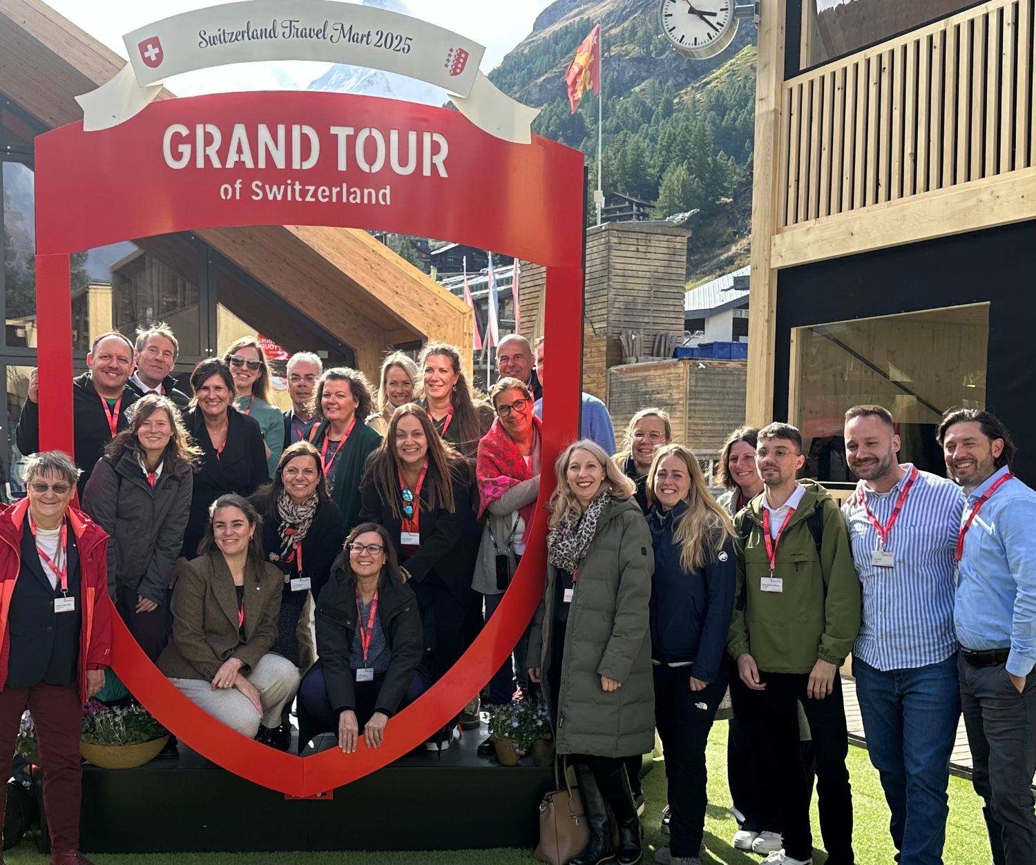Das Bild zeigt eine Gruppe von etwa 25 Menschen, die lächelnd unter einem großen roten Bogen mit der Aufschrift „GRAND TOUR of Switzerland“ stehen. Im Hintergrund sind Holzhäuser, ein Uhrenturm und Berge zu sehen. Viele Personen tragen Namensschilder und rote Bänder um den Hals, was auf eine Veranstaltung oder ein Treffen, vermutlich das „Switzerland Travel Mart 2025“, hinweist. Die Atmosphäre wirkt freundlich und entspannt, es ist Tageslicht.